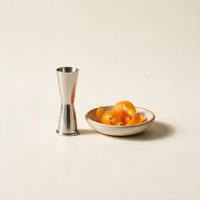 A slim polished steel jigger beside a dish of citrus peel