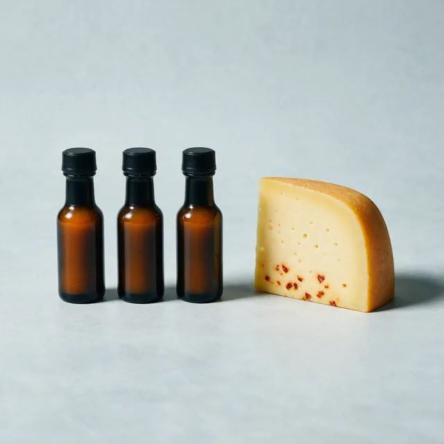 Three small bottles of hot sauce beside a wedge of hard cheese